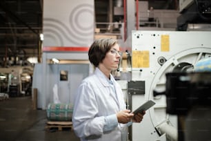 Portrait of female factory worker wearing glasses and white robe, using tablet for quality control and logistic purposes at polymer plastic manufacturing. Side view