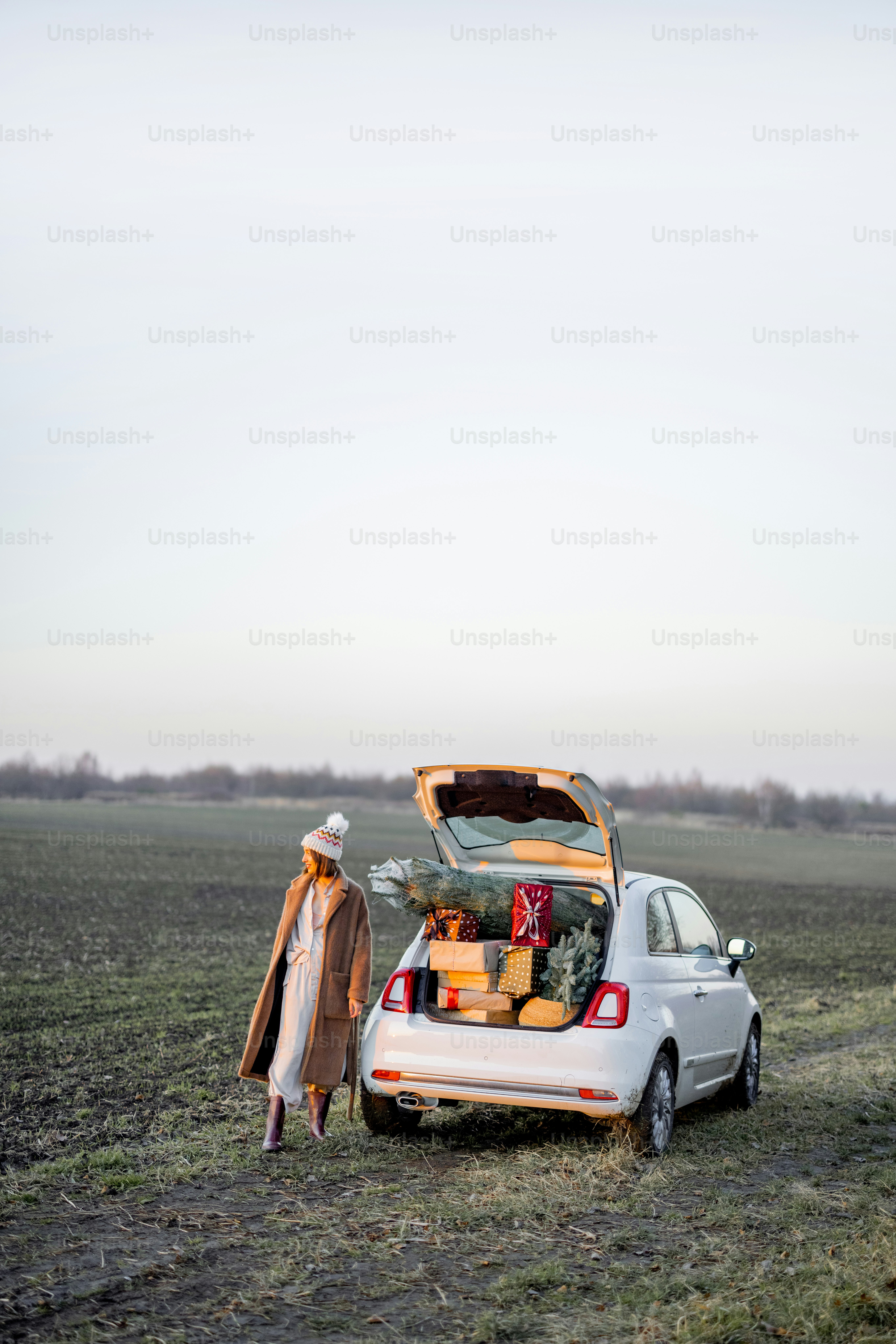 Woman in winter clothes walks near car full of giftboxes and Christmas tree on the field at sunset. Wide landscape with copy space on sky. Concept of New Year's mood