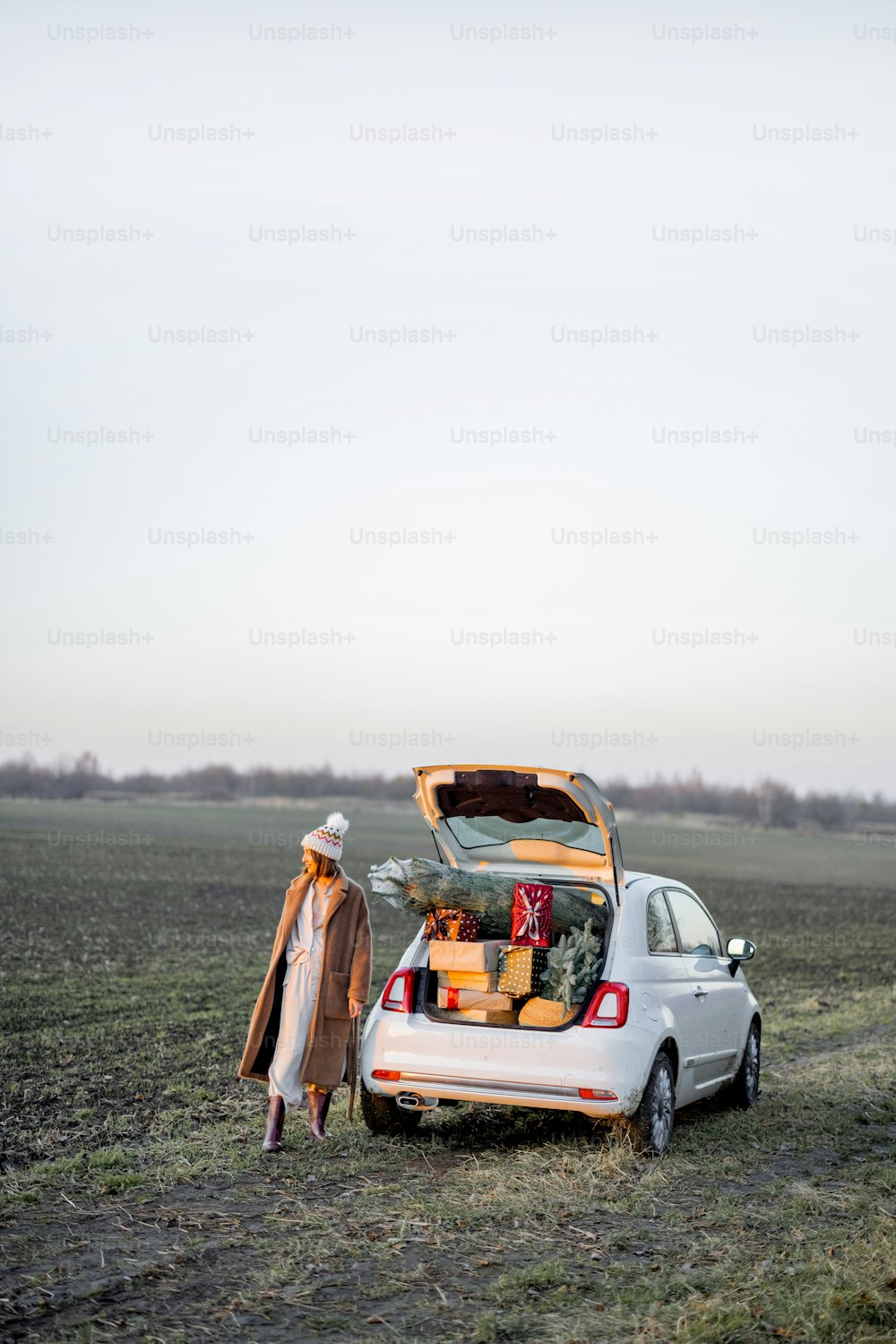 La donna in abiti invernali cammina vicino all'auto piena di scatole regalo e albero di Natale sul campo al tramonto. Ampio paesaggio con spazio di copia sul cielo. Concetto di umore di Capodanno
