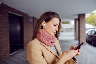 Profil d’une jeune femme à la mode en chaleur avec des écouteurs debout à l’extérieur par temps froid et tapant un message au téléphone. Internet et réseau mondial. Une femme utilisant son téléphone.