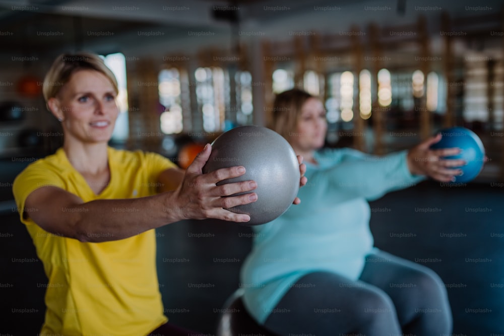 Un entrenador personal entrenando a una mujer con sobrepeso, sentada en fintess bal y haciendo ejercicio en el gimnasio.