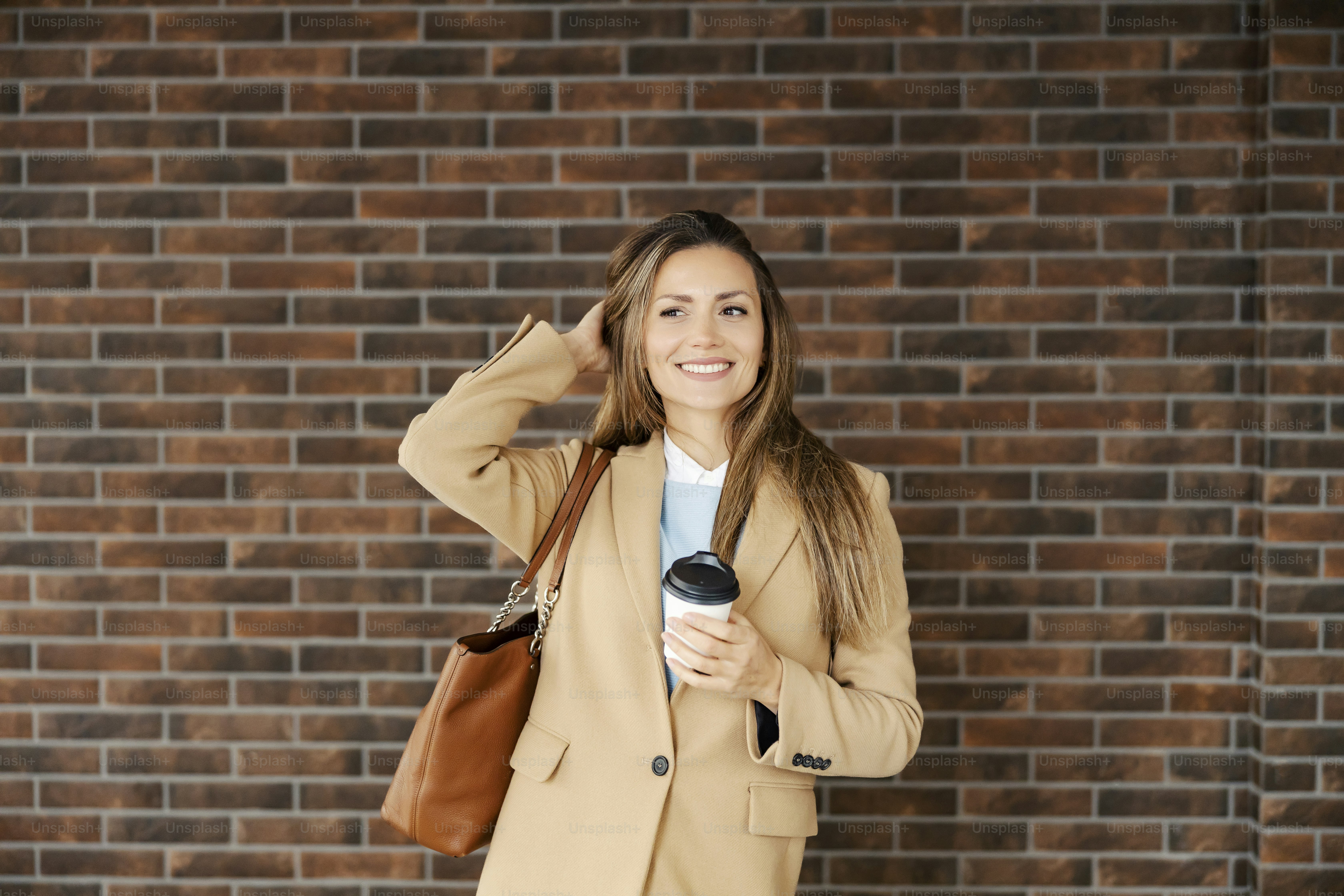 A happy woman adjusting her hair and holding takeaway coffee while standing outside. City lifestyle. A woman with coffee.