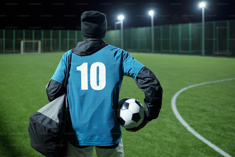 Back of football player with soccer ball and bag moving towards field while going to have night training before match