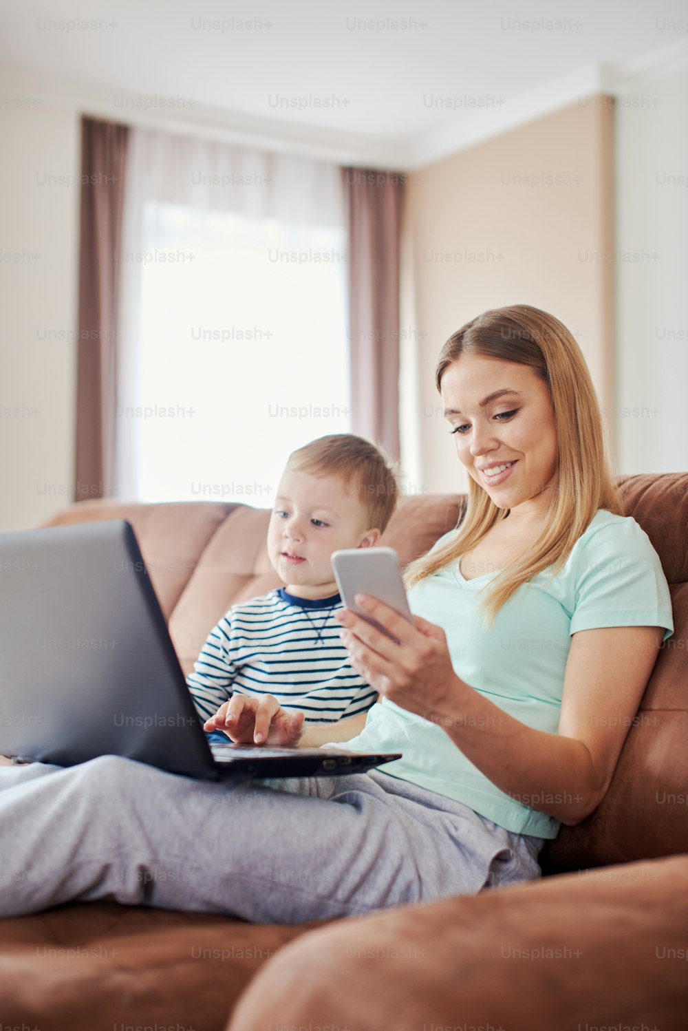Young caucasian mother with toddler son is using laptop and smart phone while sitting on the couch at home, wearing pyjamas