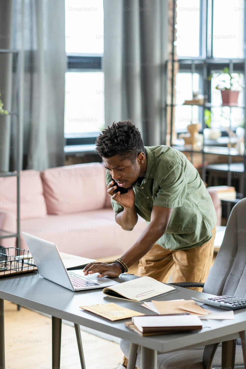 Ernsthafter junger Schwarzer, der am Tisch steht und Informationen auf dem Laptop überprüft, während er im Büro telefoniert