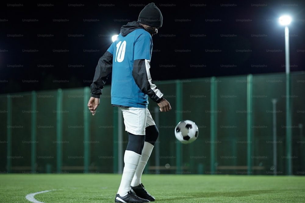 Joven negro en uniforme de fútbol profesional pateando la pelota de fútbol mientras juega en el campo rodeado de iluminaciones
