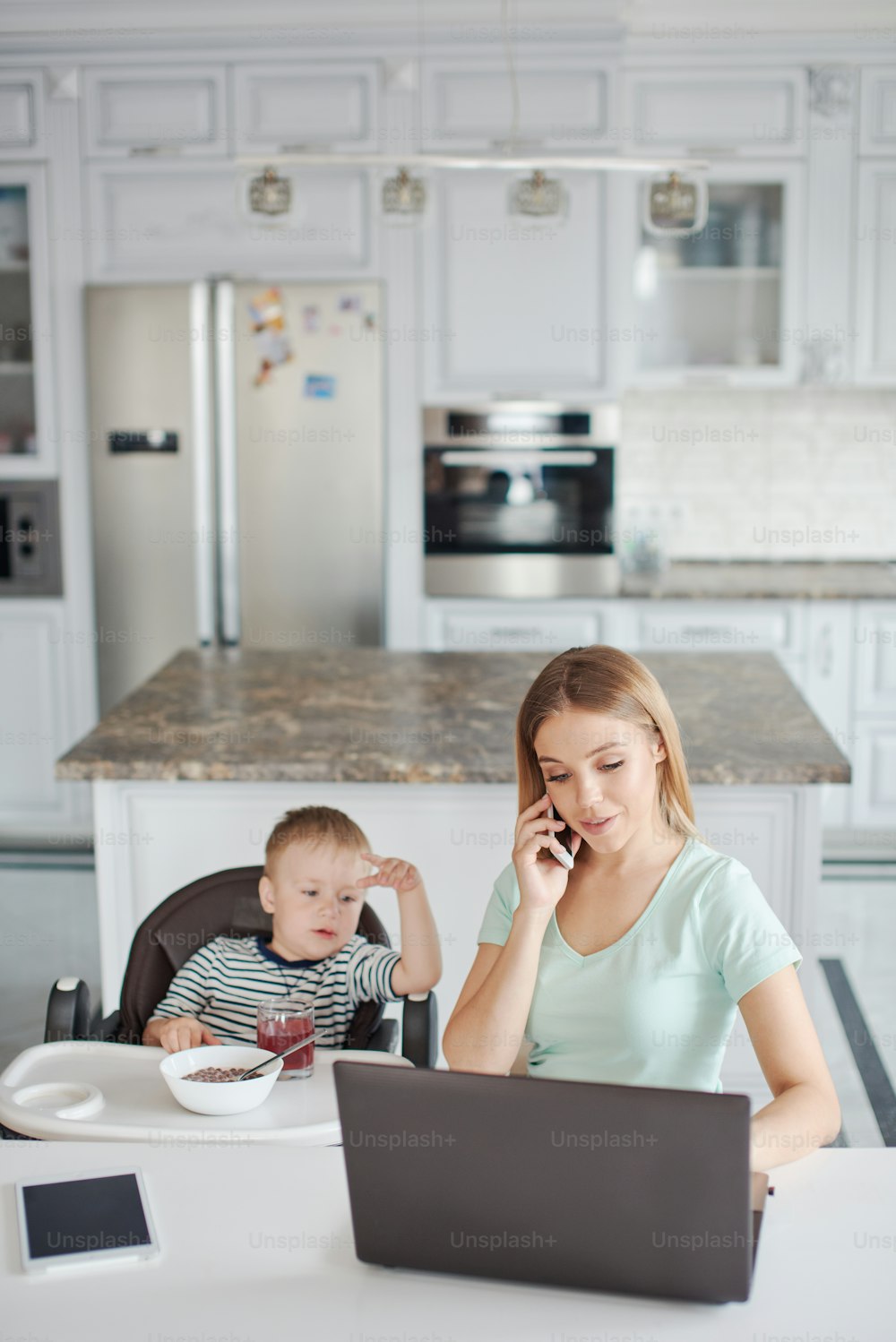 Portrait of mid age caucasian blond woman with child being busy on the phone and feeding the baby. They are at home, wearing pyjamas