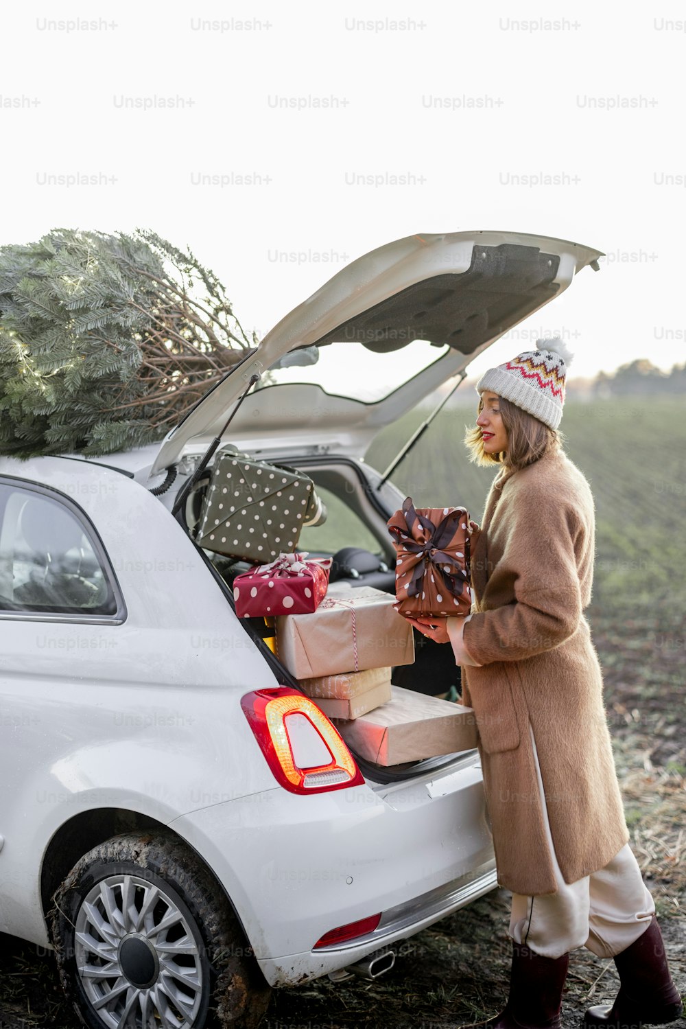 夕暮れ時の自然の屋上でクリスマスツリーを載せた車にプレゼントを詰め込む女性。年末年始の準備。クリスマスムードのアイデア。毛皮のコートと帽子をかぶった女性