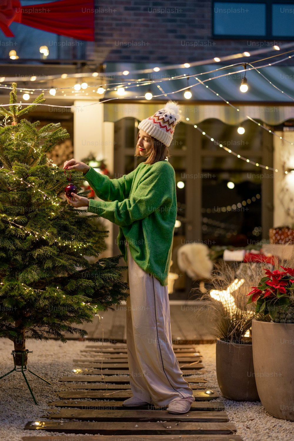 Young woman decorating Christmas tree at beautiful backyard of her country house alone. Concept of preparation for the winter holidays. Caucasian woman wearing green sweater and hat
