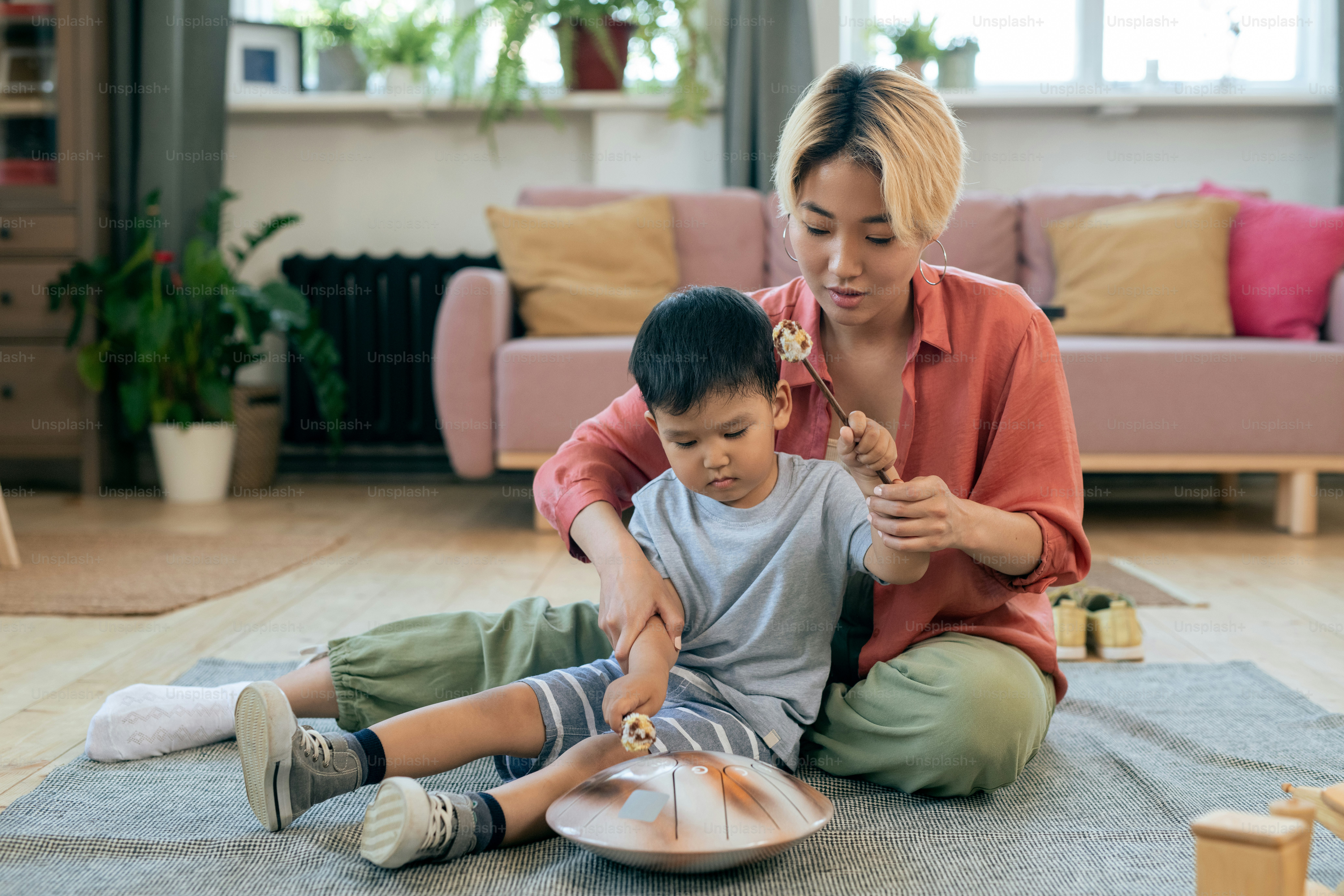 Young mother helping her cute little son hit hang drum while both sitting on the floor of living-room