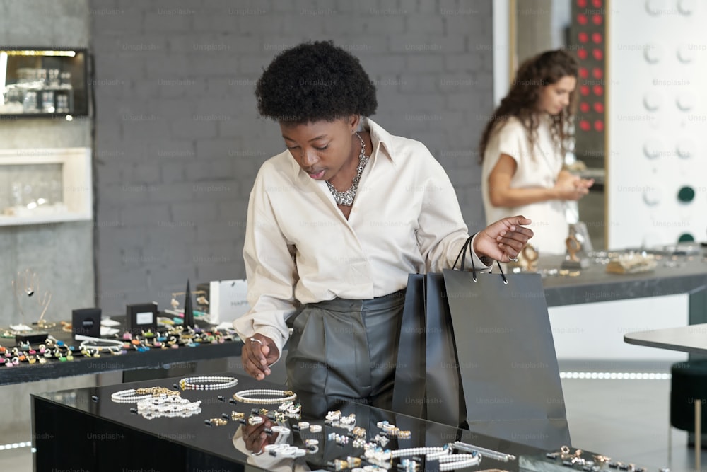 Jeune acheteuse africaine avec des sacs en papier choisissant un nouveau collier sur un grand écran tout en regardant un assortiment de bijoux