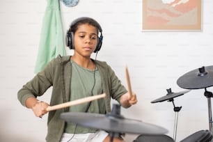 Linda colegiala birracial en auriculares y ropa casual tocando la batería mientras entrena en el entorno del hogar