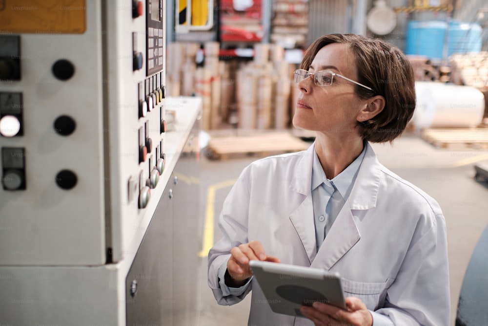 Portrait of female factory worker wearing glasses and white robe, using tablet for quality control and logistic purposes at polymer plastic manufacturing