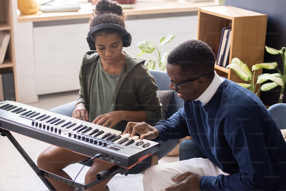 Cute biracial girl in headphones looking at music teacher pressing keys of keyboard during lesson at home