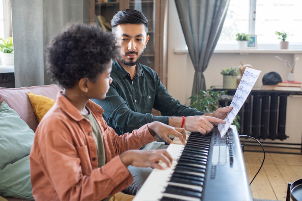 Junger Musiklehrer, der auf ein Blatt mit Noten zeigt, während er den niedlichen, fleißigen Schuljungen betrachtet, der Klavier spielt, Keyboard spielt