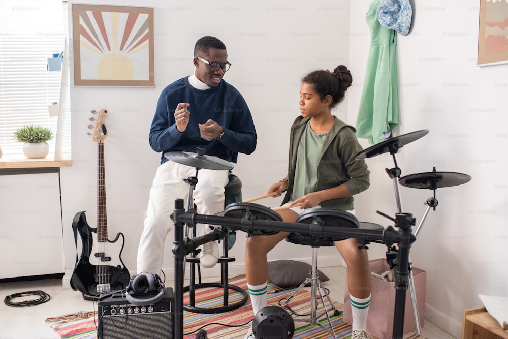 Cute diligent schoolgirl hitting drums with drumsticks at lesson of music while teacher consulting her