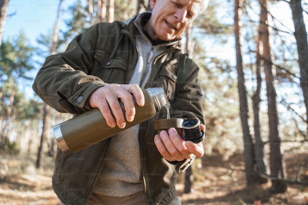 Niedrige Winkelansicht des glücklichen alten Mannes, der heißes Getränk aus der Thermoskanne in die Tasse gießt, während er sich nach einem Spaziergang im Wald ausruht