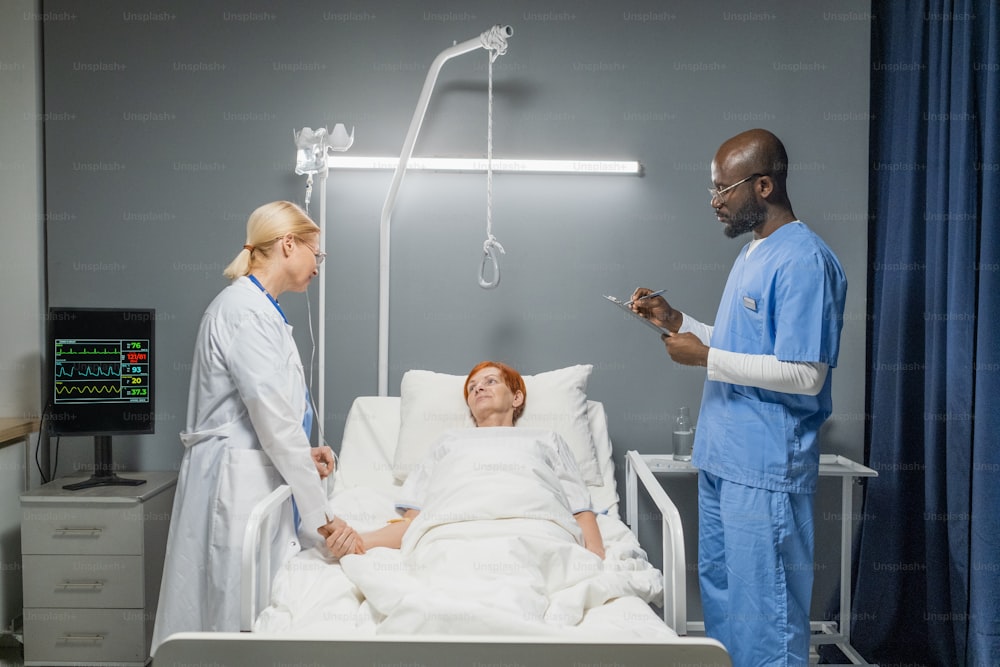 Médico africano en uniforme tomando notas en la tarjeta médica mientras la enfermera habla con una anciana mientras está acostada en la cama en la sala del hospital