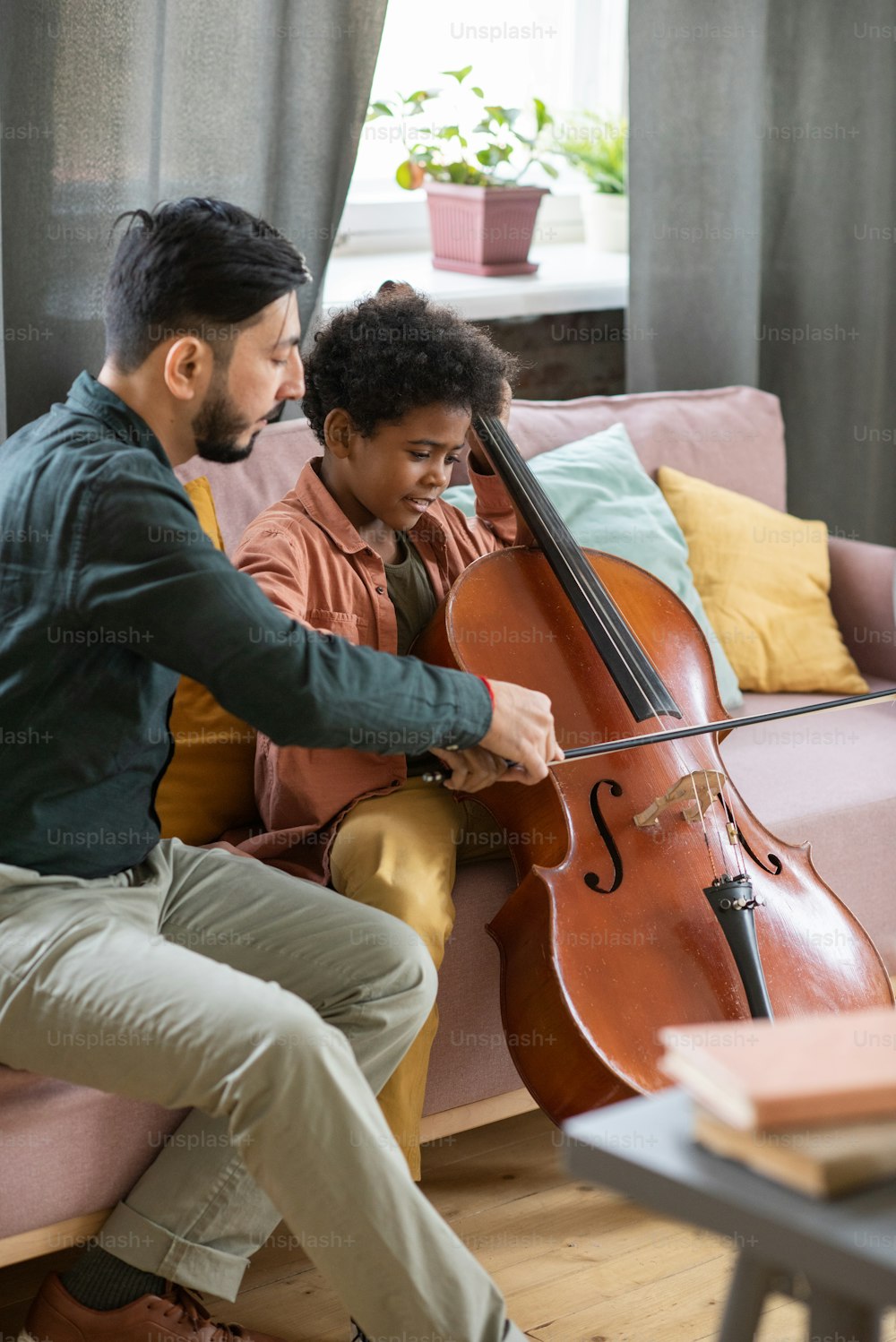 Joven profesor de música que ayuda a un lindo colegial que toca el violonchelo mientras ambos están sentados en el sofá en el entorno del hogar