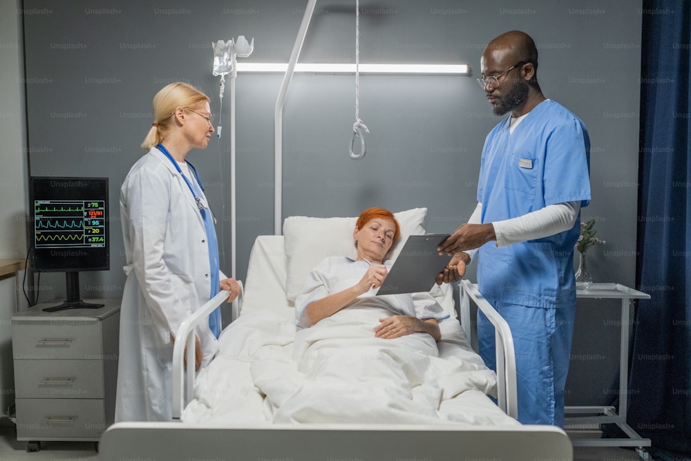 Ältere Frau unterschreibt eine Vereinbarung über die Operationsabgabe durch den Anästhesisten und Arzt, die in der Nähe des Bettes auf der Krankenstation stehen