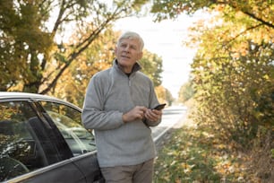 Hombre maduro concentrado que mira hacia otro lado con expresión pensativa mientras está parado cerca de su automóvil en el bosque y usa un teléfono inteligente. Cálido día de otoño