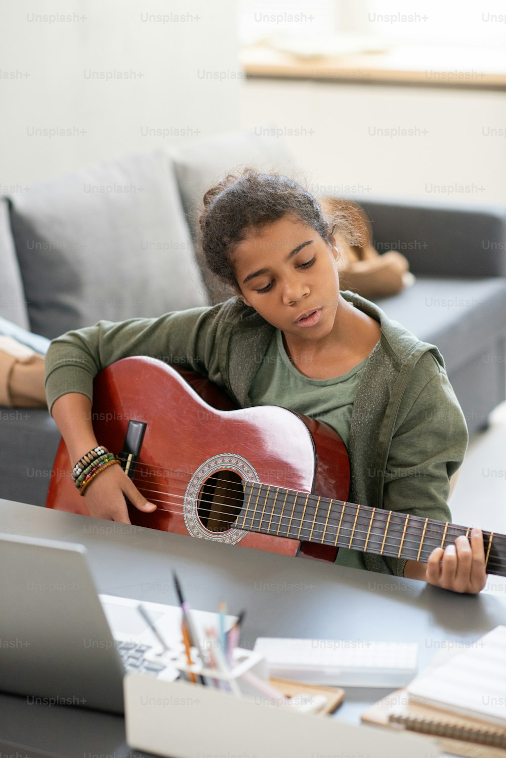 Linda preadolescente con guitarra acústica tocando frente a la computadora portátil durante la lección en línea en el entorno del hogar