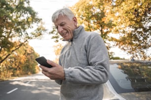 Low angle view of the grey haired man wearing warm clothes holding his smartphone and looking at the screen while standing at the road near the forest