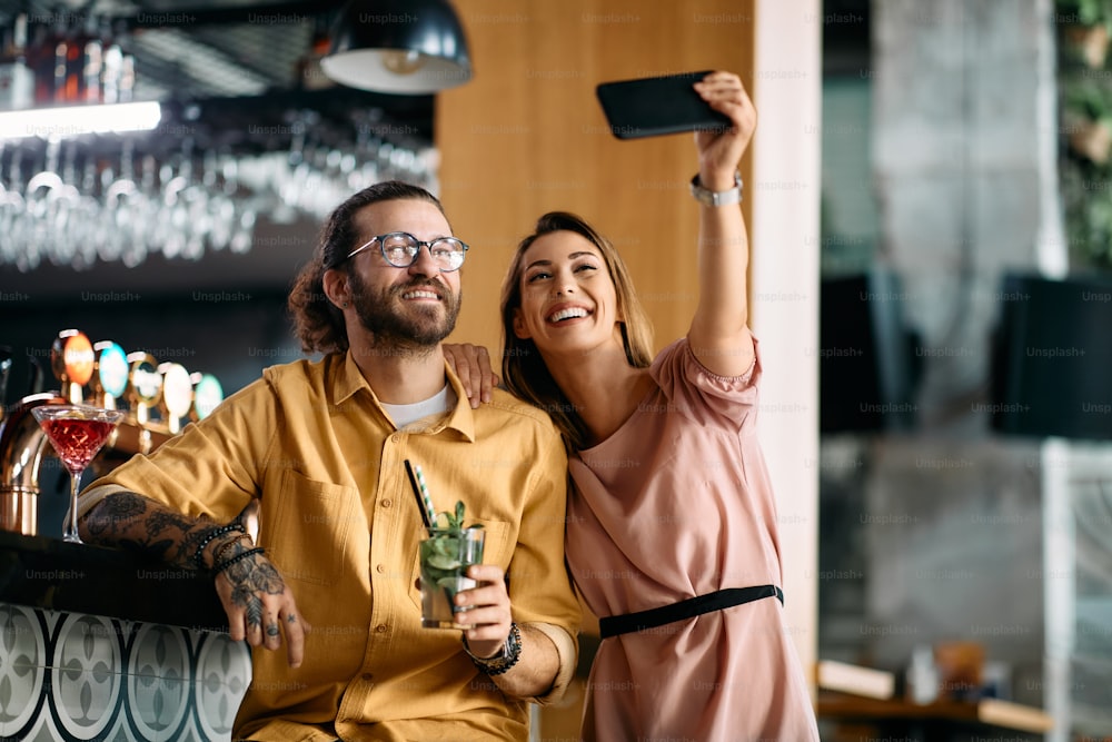 Happy couple using smart phone and taking selfie while drinking cocktails in a bar.