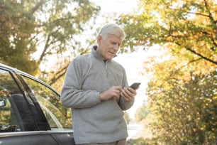 Vista de ángulo bajo del anciano caucásico buscando algo en el teléfono inteligente mientras está parado cerca de su automóvil y se prepara para caminar por el bosque