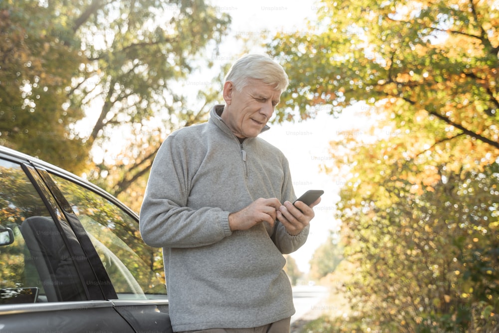 Vista dal basso dell'anziano caucasico che cerca qualcosa allo smartphone mentre si trova vicino alla sua auto e si prepara per la passeggiata nella foresta
