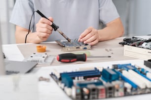 Hände eines jungen Mechanikers, der winzige Details auf dem Motherboard mit Lötkolben fixiert