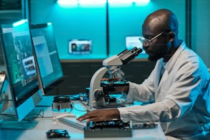 Joven investigador africano con microscopio presionando la tecla del teclado de la computadora durante el trabajo