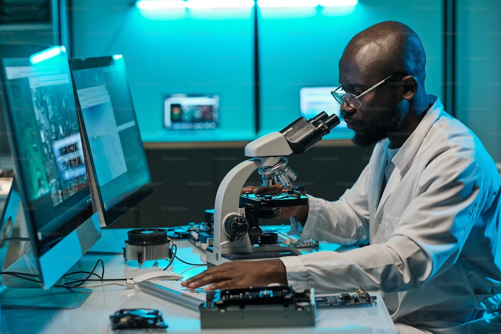 Joven investigador africano con microscopio presionando la tecla del teclado de la computadora durante el trabajo