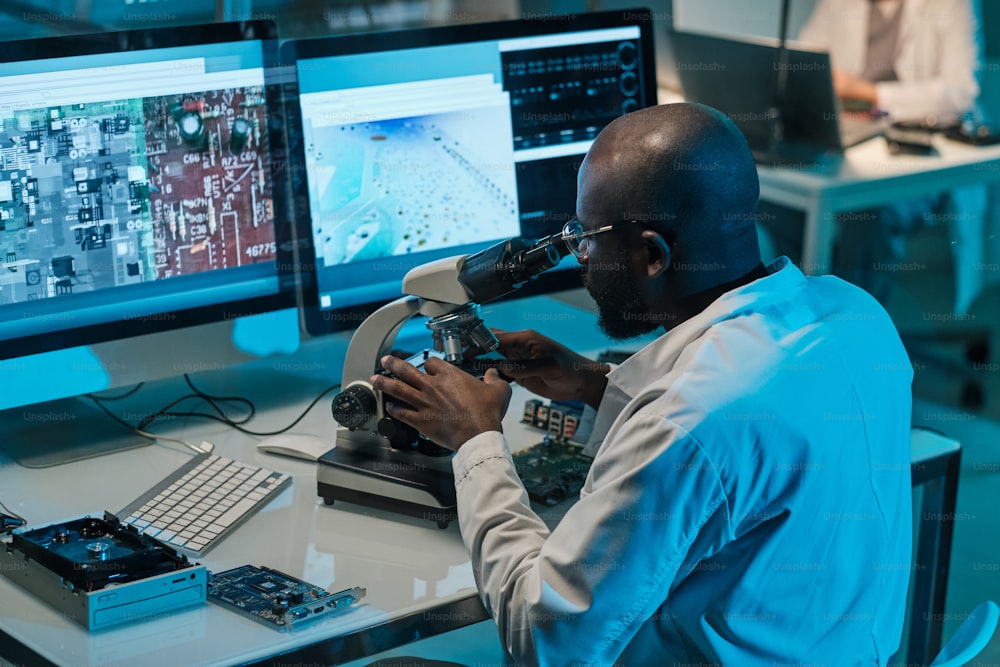 Contemporary young African scientist with microscope working in front of computer monitors