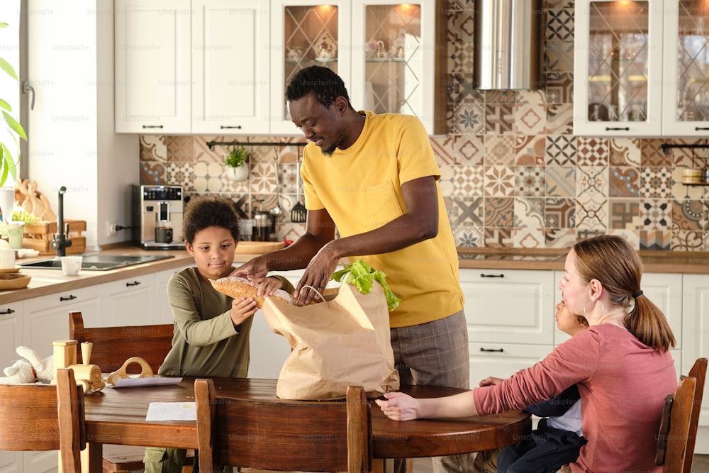 Lindo niño birracial que ayuda a su padre a sacar pan fresco comprado y otros productos de una bolsa de papel junto a la mesa de la cocina