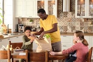 Cute little biracial boy helping his father take out bought fresh bread and other products out of paperbag by kitchen table