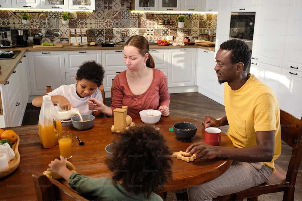 Contemporary interracial family of four having breakfast while sitting by wooden kitchen table