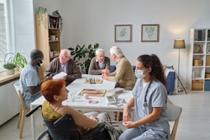 Nurse talking to elderly woman during medical exam with other senior people in the background in nursing home