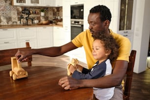 Young African man in casualwear playing wooden toys with his cute little son while sitting by table in the kitchen