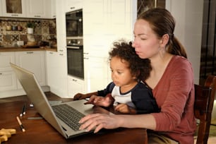 Young busy woman holding her cute little son while sitting by wooden kitchen table in front of laptop and networking
