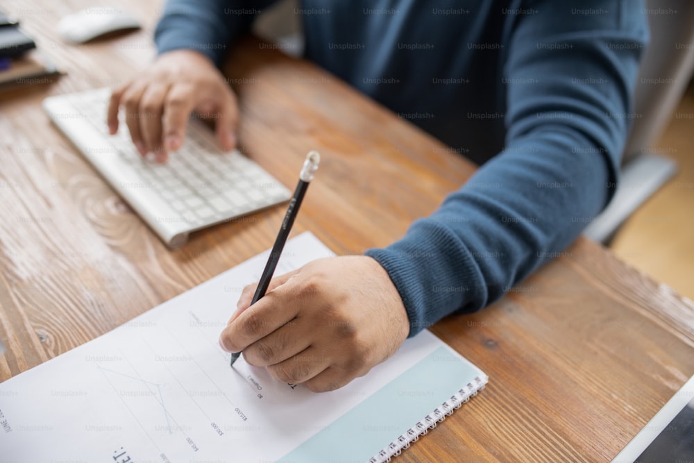 Hand eines männlichen Lehrers oder Ökonomen mit Stift, der auf Finanzdokumente zeigt, während er am Tisch sitzt und arbeitet
