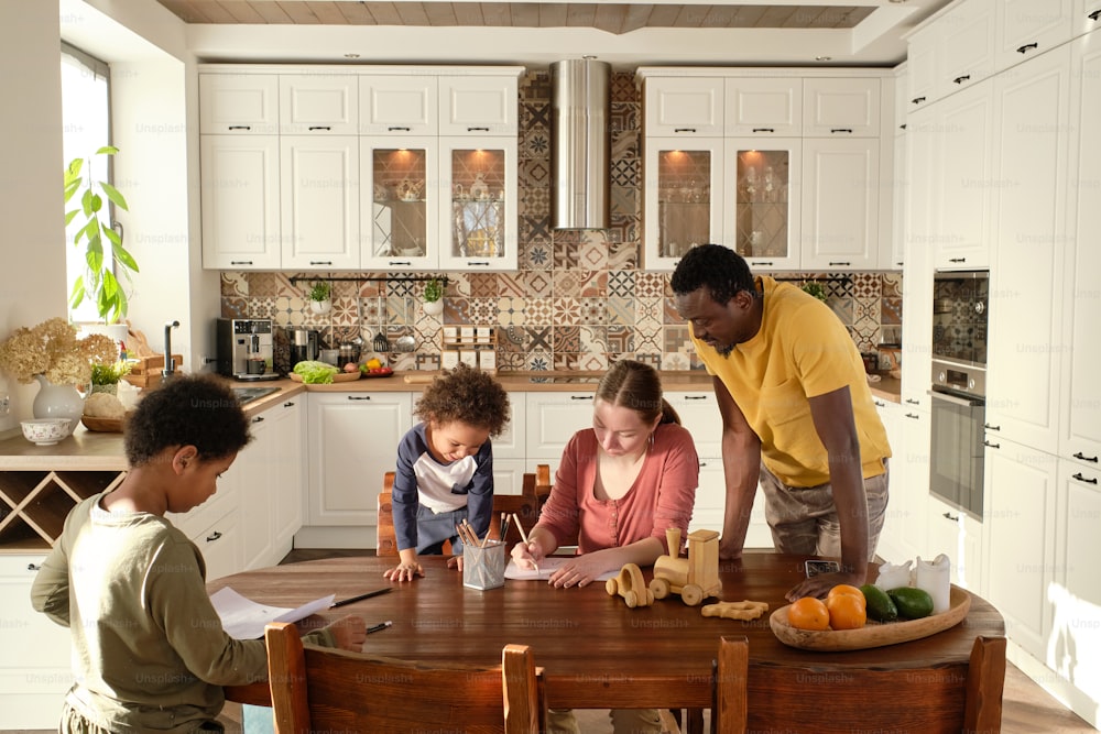 Contemporary young family of parents and two little sons drawing together while gathered by wooden kitchen table