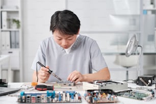 Giovane tecnico cinese femminile in uniforme che ripara il circuito stampato con microprocessore mentre si siede sul posto di lavoro