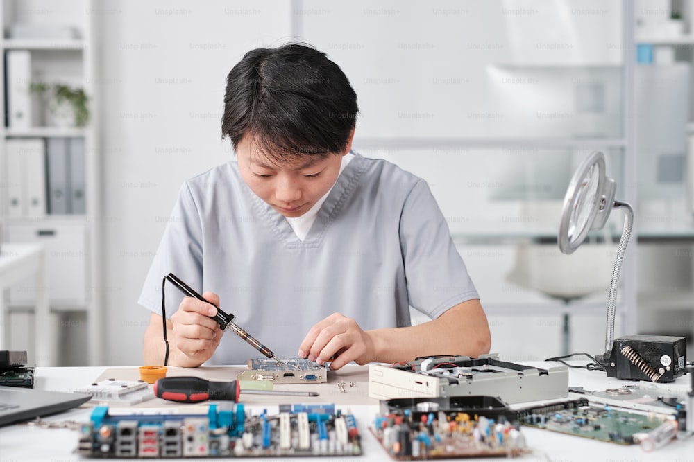 Joven técnica china en uniforme que repara la placa de circuito con microprocesador mientras está sentada en el lugar de trabajo