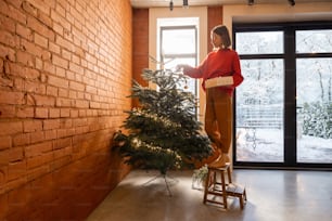 Young woman decorating Christmas tree at home. Concept of home comfort and preparation for a winter holidays. Girl wearing red sweater, living room with windows overlooking on snowy garden