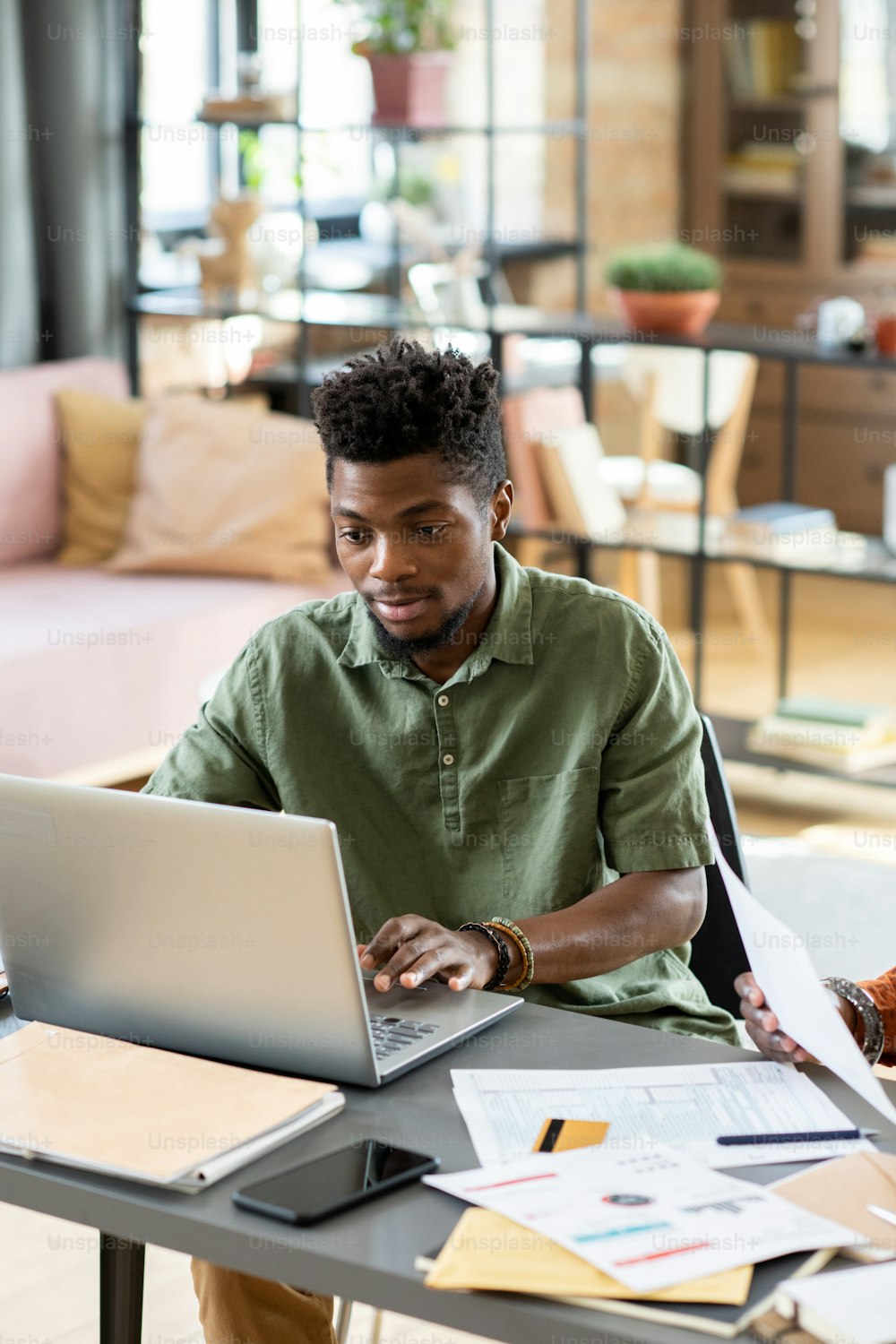 Junger Geschäftsmann afrikanischer Abstammung schaut auf das Laptop-Display beim Networking am Tisch in häuslicher Umgebung