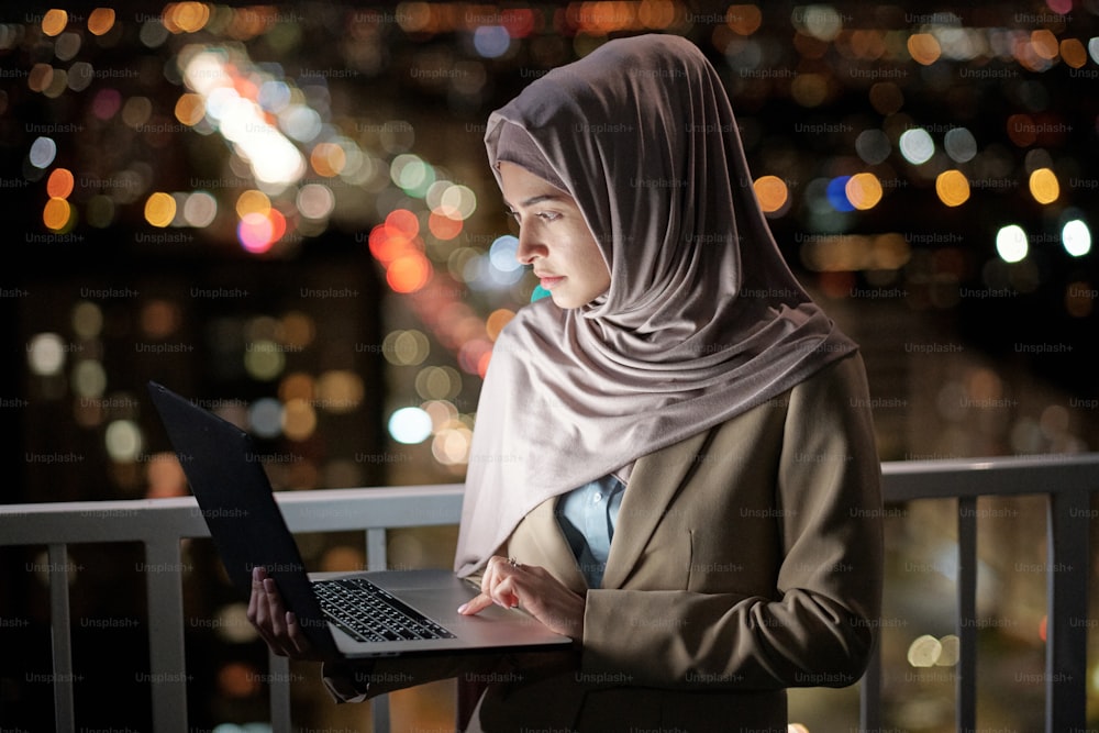 Serious young businesswoman with laptop looking at its screen while networking against urban environment at night