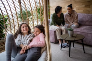 Hermanitas felices sentadas en mimbre de ratán cuelgan silla en el interior del invernadero, con la madre y la abuela en el fondo.