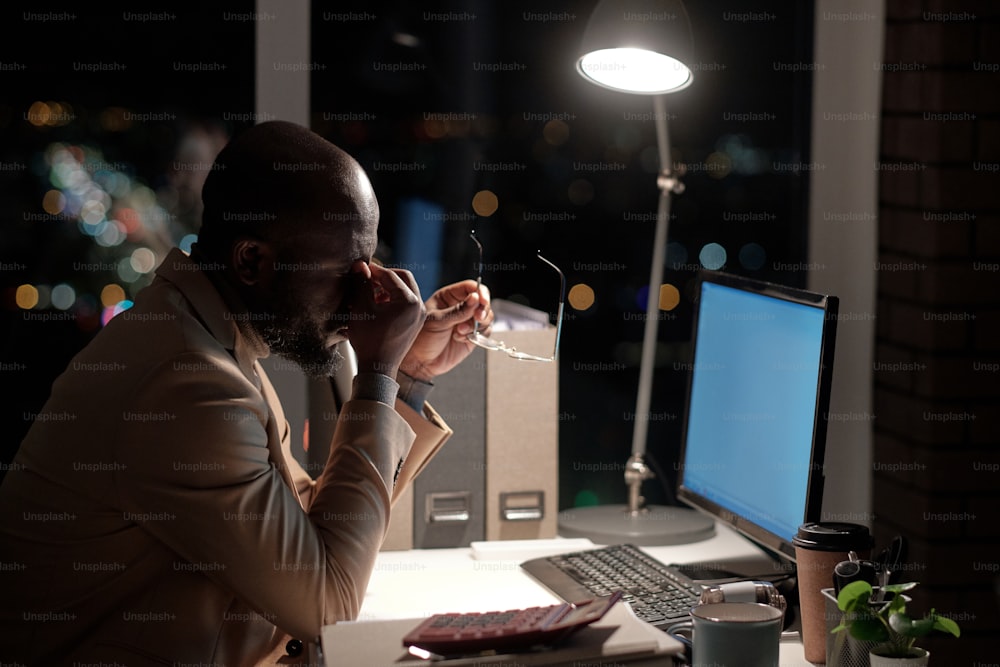 Young tired businessman touching his face while working in front of computer screen in office late at night