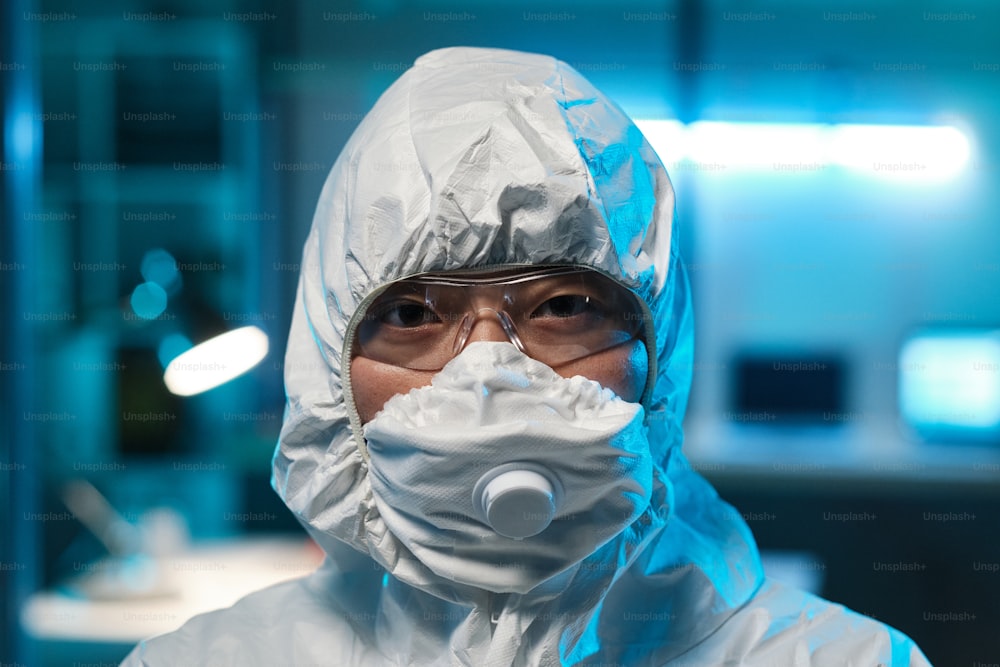Head of young Asian female laboratory worker or scientist in protective eyeglasses, respirator and coveralls